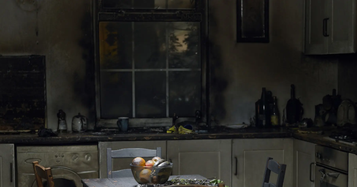 Kitchen after experiencing fire damage, showing a window, burn marks on the wall, and various items on the table, counters, and in the sink